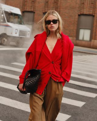 A woman wears a red cardigan, brown trousers, and black bag with a red sweater over her shoulders.