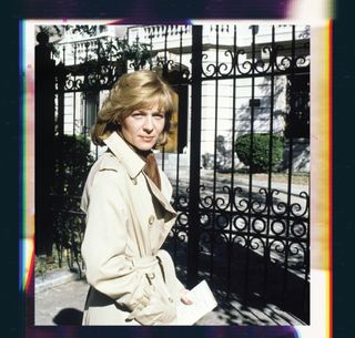 Woman with blonde hair in a khaki trench coat standing in front of a fence.