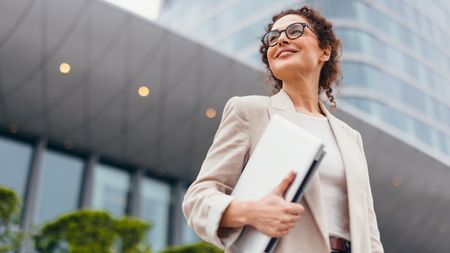 A businesswoman outside an office building with a laptop under her arm.