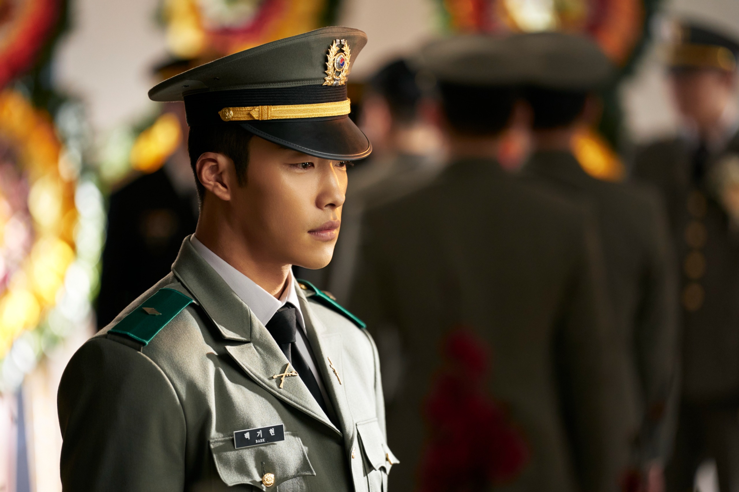A Korean man in a 1970 military uniform (Woo Do-hwan as Baek Kihyun) stands in front of several soldiers and floral wreaths, in Hulu's 'Made in Korea.'