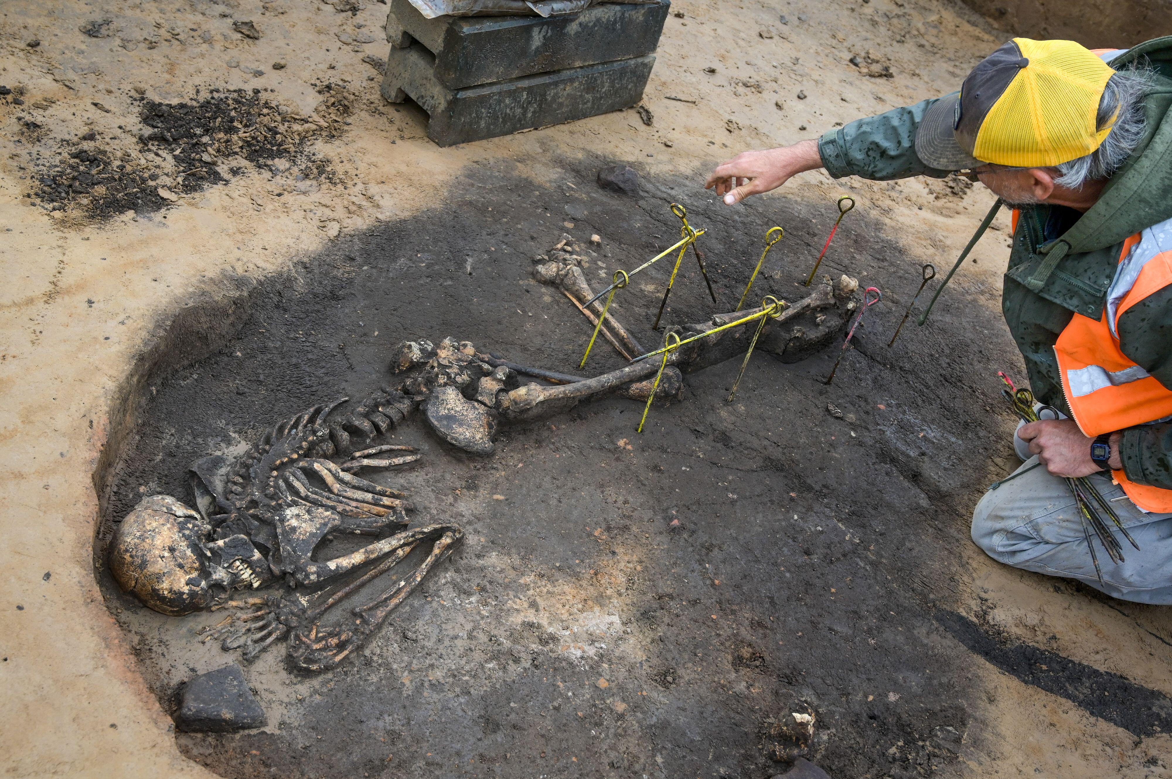 a light-skinned man in high-viz gear excavates a human skeleton from an archaeological site
