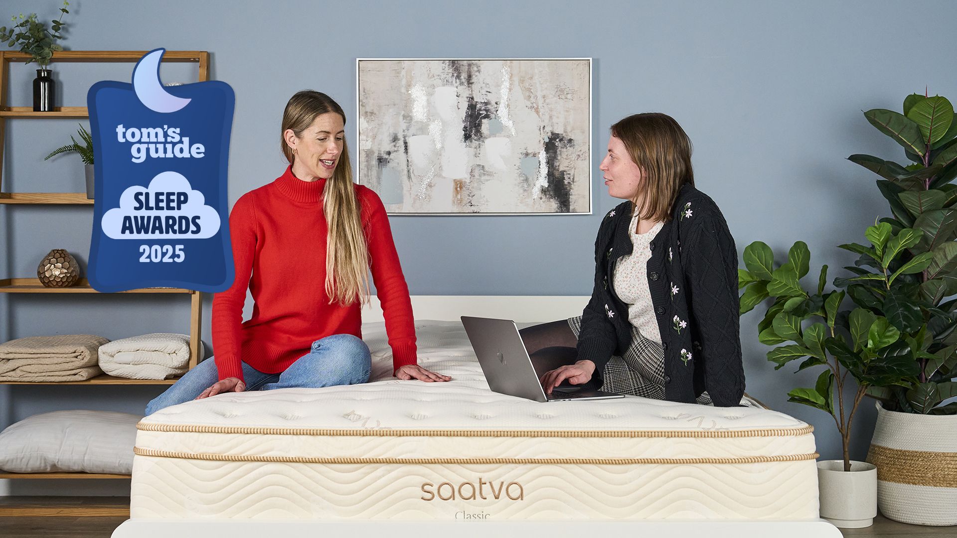 The image shows two women sitting on top of the cream and gold Saatva Classic mattress. One is examining the surface of the mattress while the other is making notes on a laptop. There is a Tom's Guide Sleep Awards 2025 badge laid over the top of the image. 