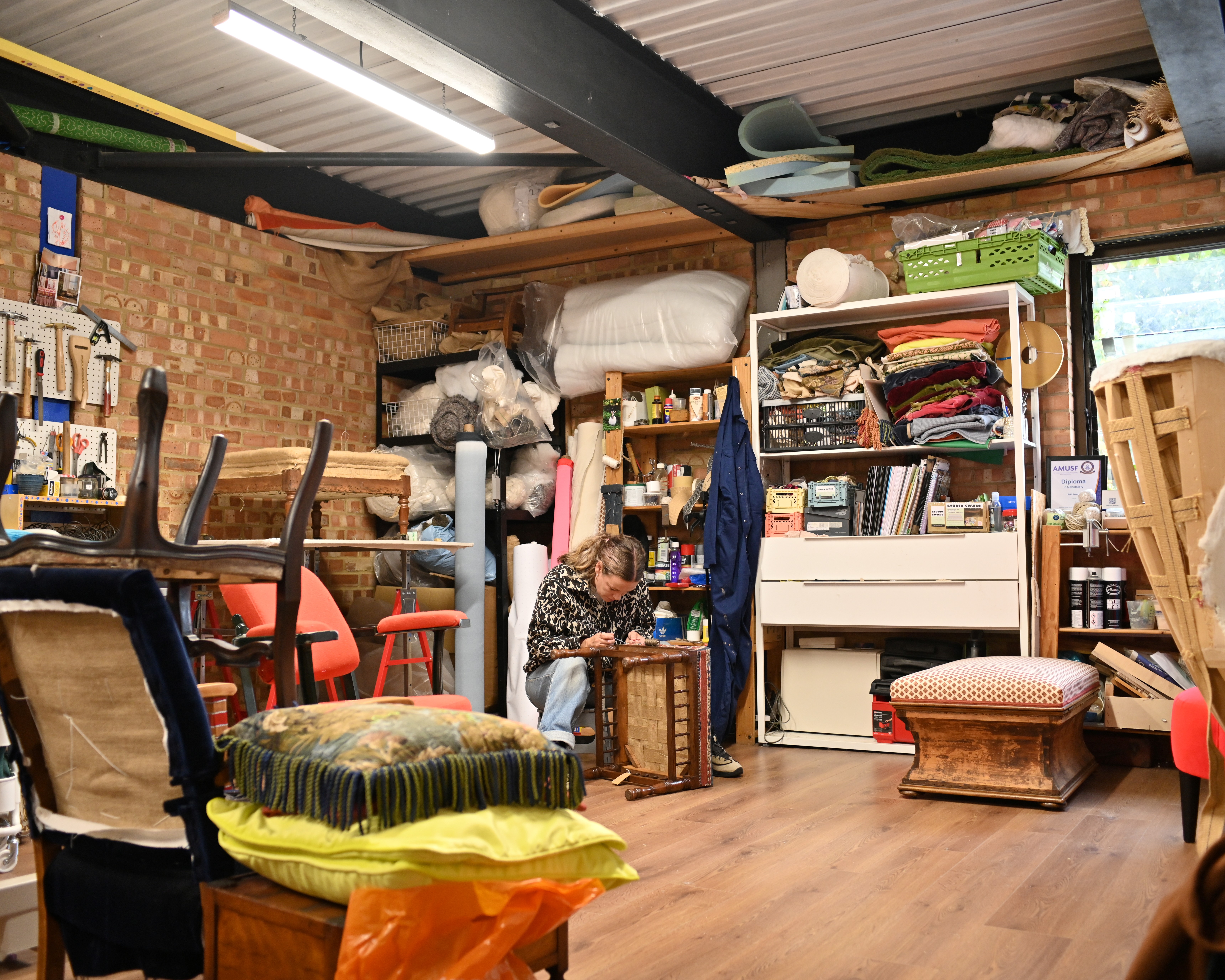 An female upholsterer photographed at work, up close and in portraits, in her studio, filled with wooden furniture, colorful fabric rolls, and mockup designs, and illuminated by bright light.