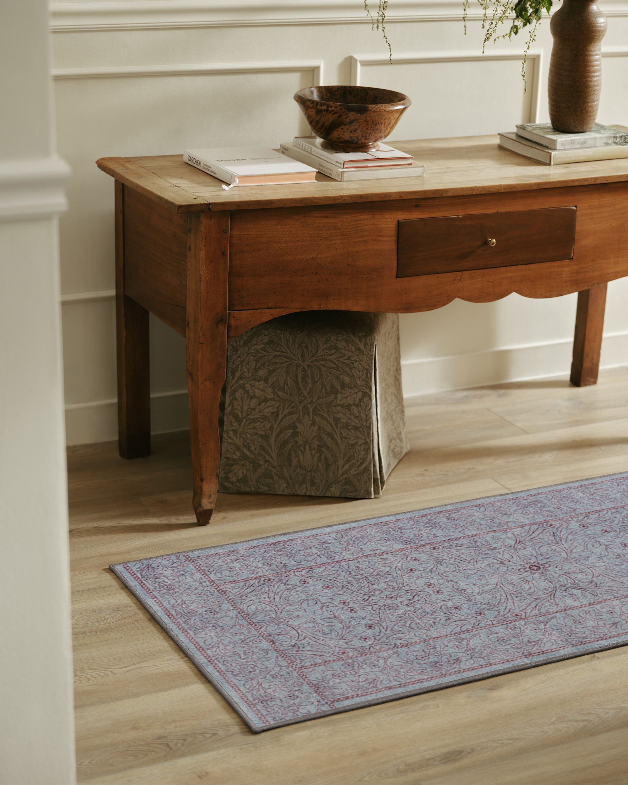 A blue and burgundy runner rug in a white entryway with a wooden console table with books and catch-alls on it.