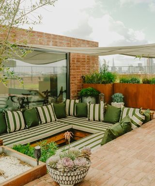 Built-in outdoor banquette seating with striped green and cream cushions, surrounded by lush plants and warm brickwork