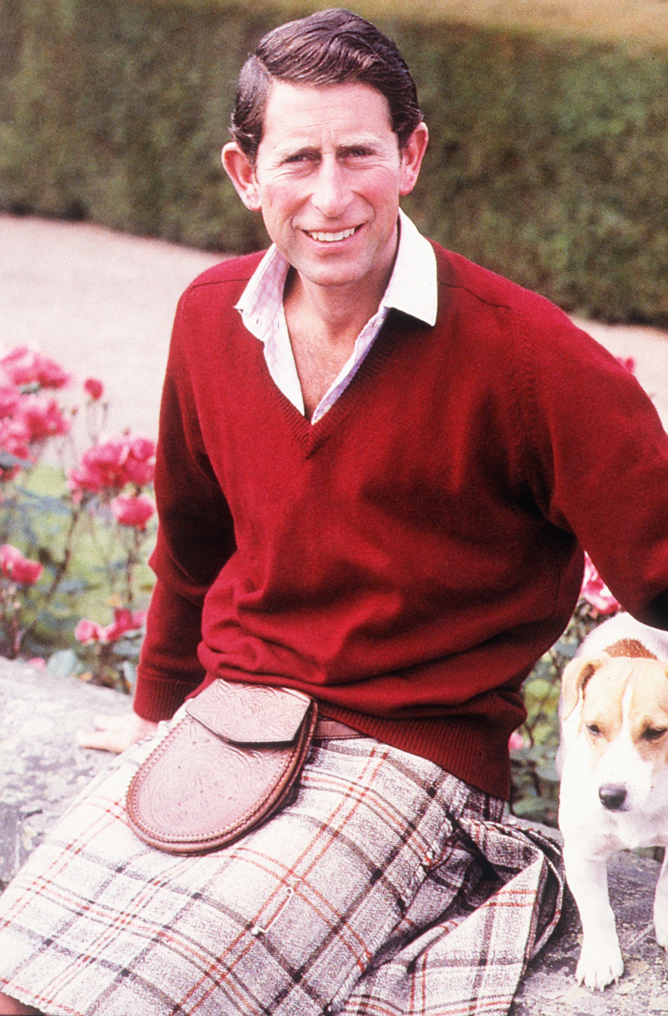 Prince Charles wearing a kilt and red sweater smiling with his jack russell Tigga