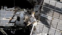 Two spacesuited cosmonauts are seen working outside a large space station set against the blackness of space.