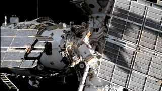 Two spacesuited cosmonauts are seen working outside a large space station set against the blackness of space.