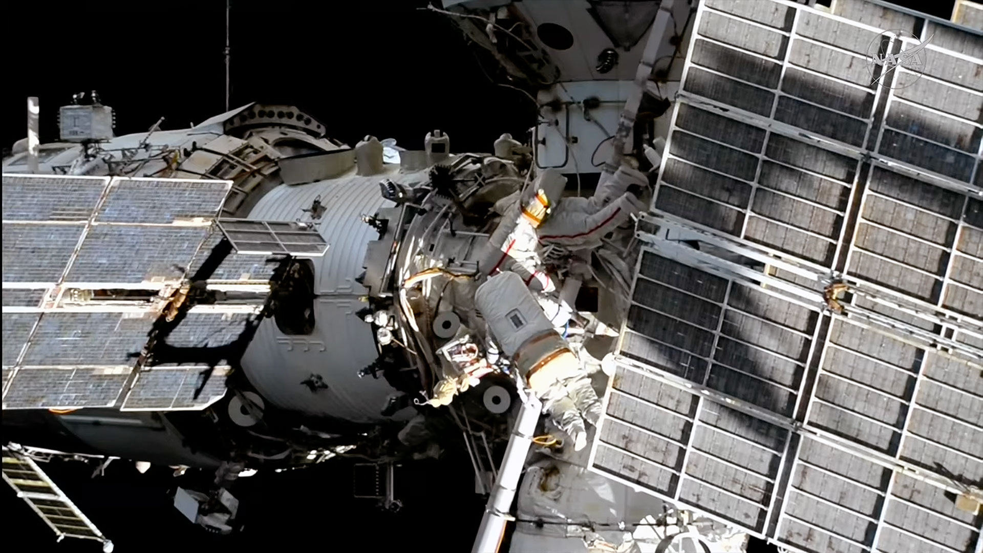 Two spacesuited cosmonauts are seen working outside a large space station set against the blackness of space.