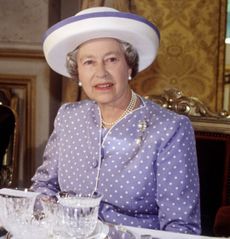 Queen Elizabeth sitting at a dinner table wearing a purple polka dot dress and white hat
