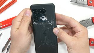 A screenshot from JerryRigEverything's durability test of the OnePlus 10 Pro, showing the phone's damage from a bend test. The back of the phone has significant cracks down the center and across the width.