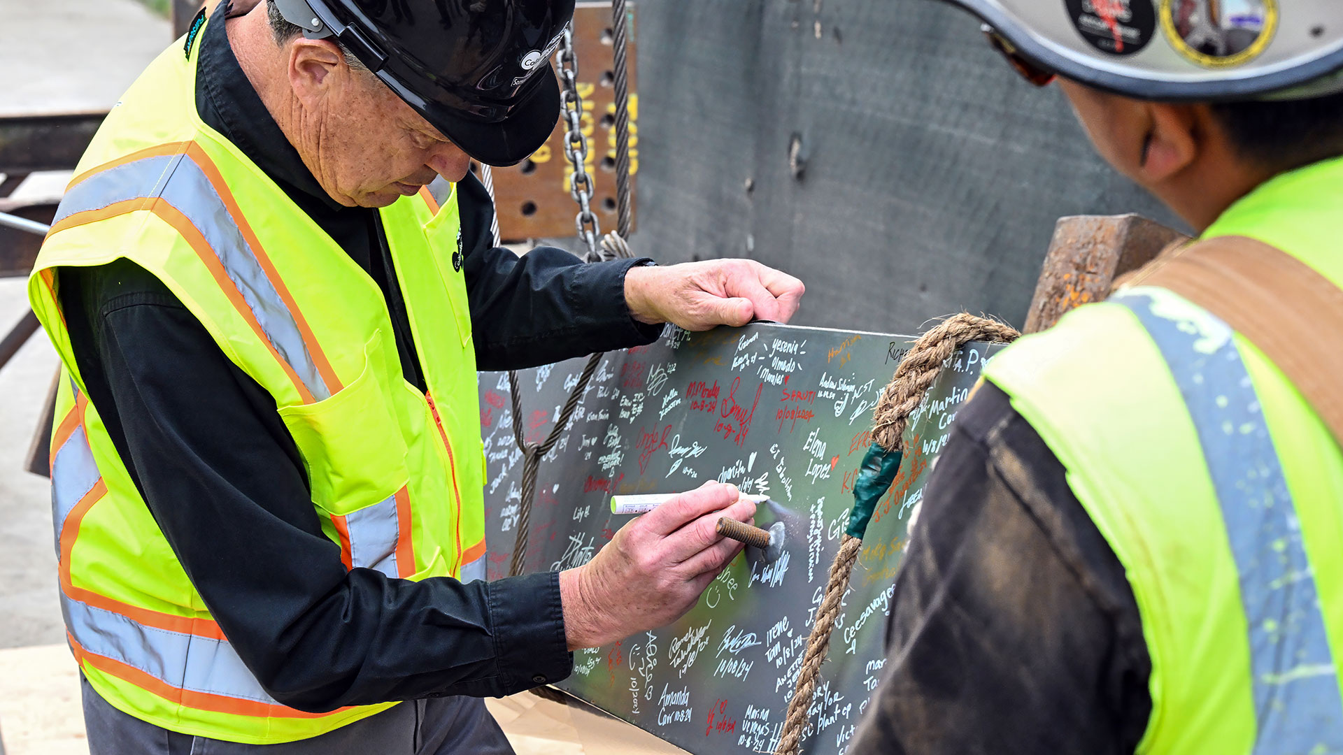 two people in hardhats and reflective yellow-green vests sign a steel beam with a marker