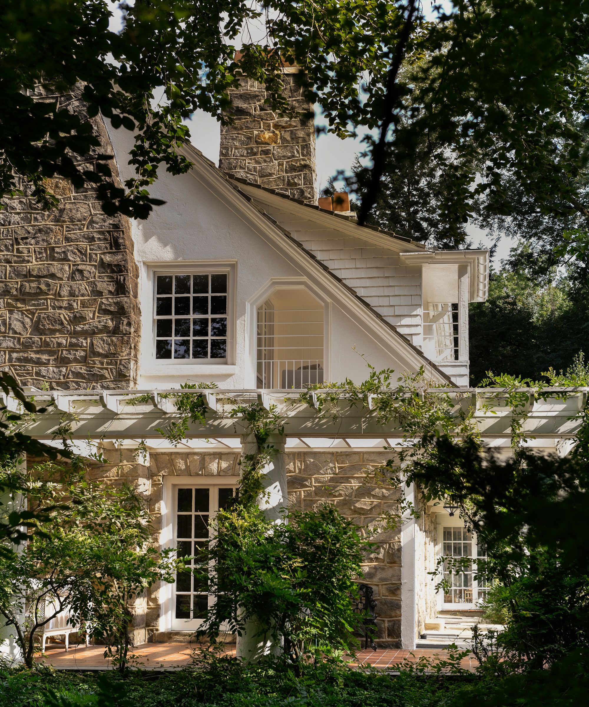 A traditional home exterior with exposed stone, white paint, and traditional-style windows, surrounded by greenery from trees.