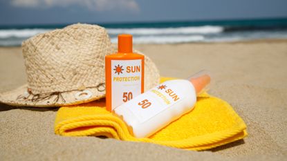 Two bottles of sunscreen on top of a yellow towel and next to a straw hat on a sandy beach .