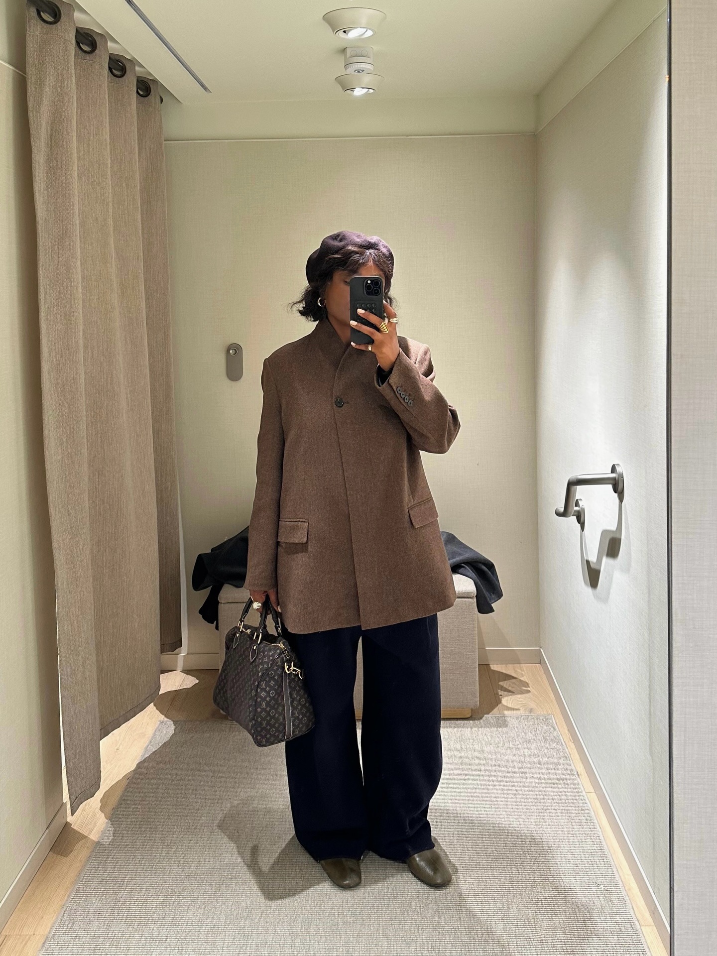 A woman taking a mirror selfie in a changing room, wearing a smart brown coat with a bob hairstyle