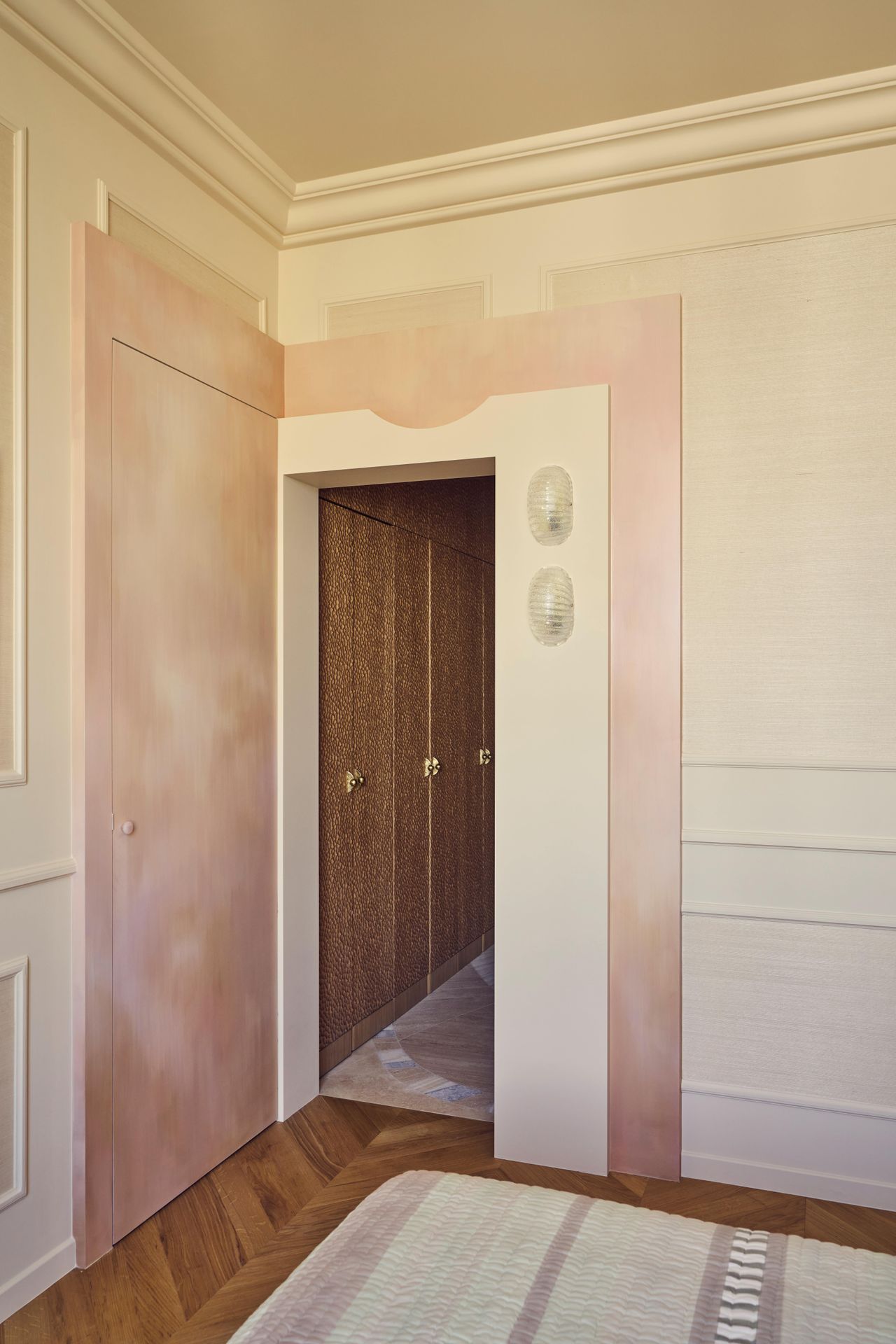 Batiik Studio-designed master bedroom furnished with mostly pastel pink and cream accents, which feel warm against the herringbone wooden floors. There is an oversized heardboard panel overhead, taking up the better portion of the wall.