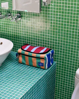 A travel kit in green, orange, red, and white blue lines sits atop a tiled bath sink in green and seafoam tones.