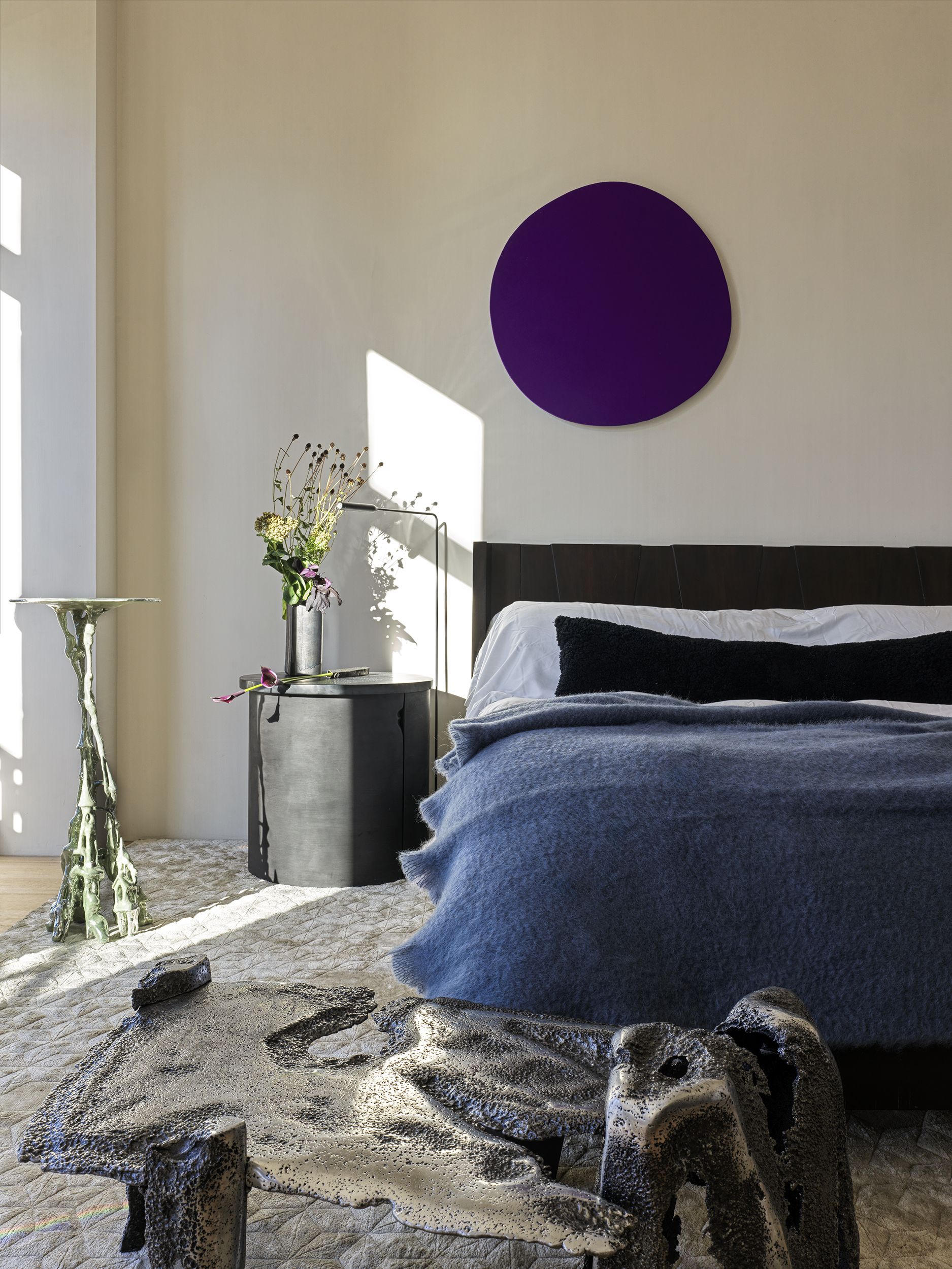 A bedroom with white walls, a black headboard and bedside with a large gray rug underneath the bed, and sculptural pieces around it and a round purple artwork above the bed