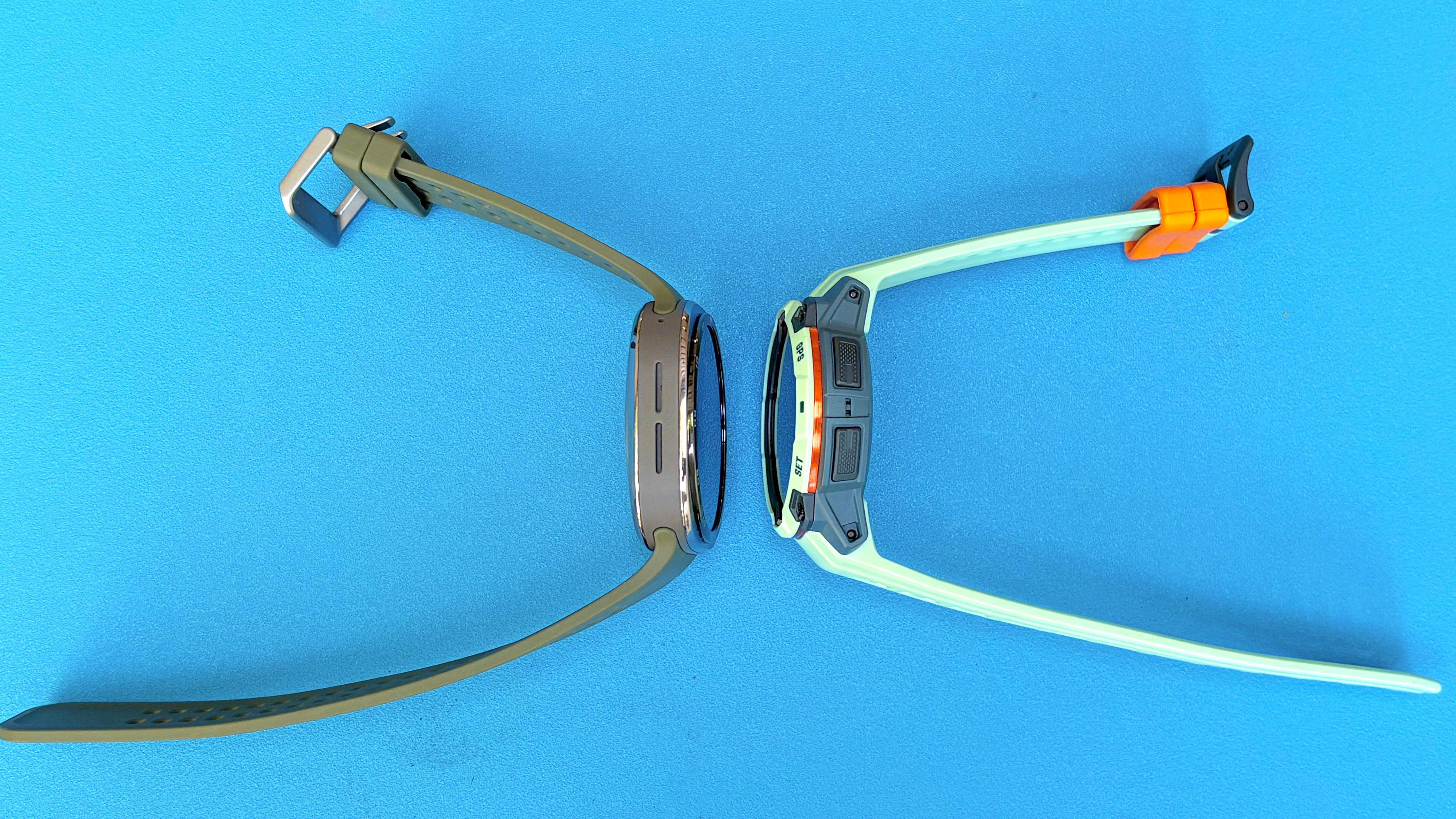 Close-up of the Samsung Galaxy Watch Ultra 2 next to the Garmin Instinct 3 against a bright blue background