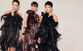Three models can be seen wearing oversized, curly dresses in metallic black, pink and blue.