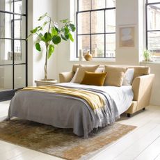 A yellow sofa bed dressed with white bed linen in a room with a large window and potted plant