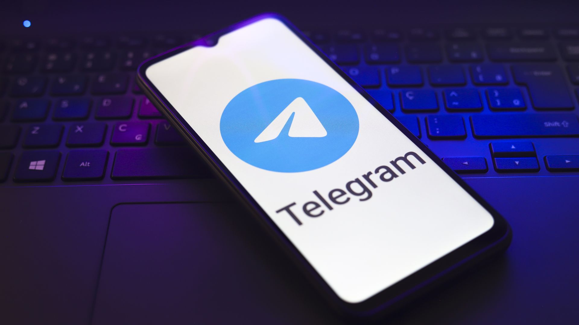 The Telegram logo appears on the screen of a smartphone that rests on top of a laptop keyboard