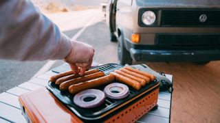 Person cooking hotdogs and onions on a camping stove
