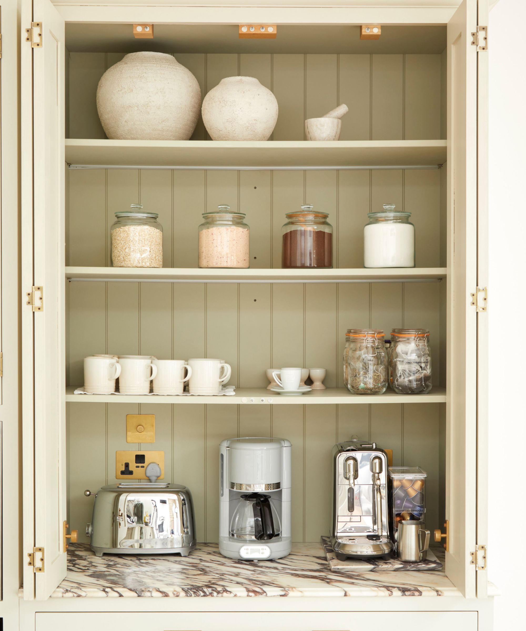 A classic kitchen cupboard transformed into a chic drinks and breakfast hub. Cream shelving displays ceramics and pantry jars, while a marble-effect countertop houses a toaster, coffee maker and espresso machine against soft, neutral cabinetry.