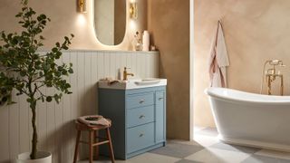 A luxurious bathroom featuring a freestanding brass-accented bath, blue vanity, tongue-and-groove panelling, a backlit mirror, warm plastered walls, and patterned floor tiles for timeless elegance.