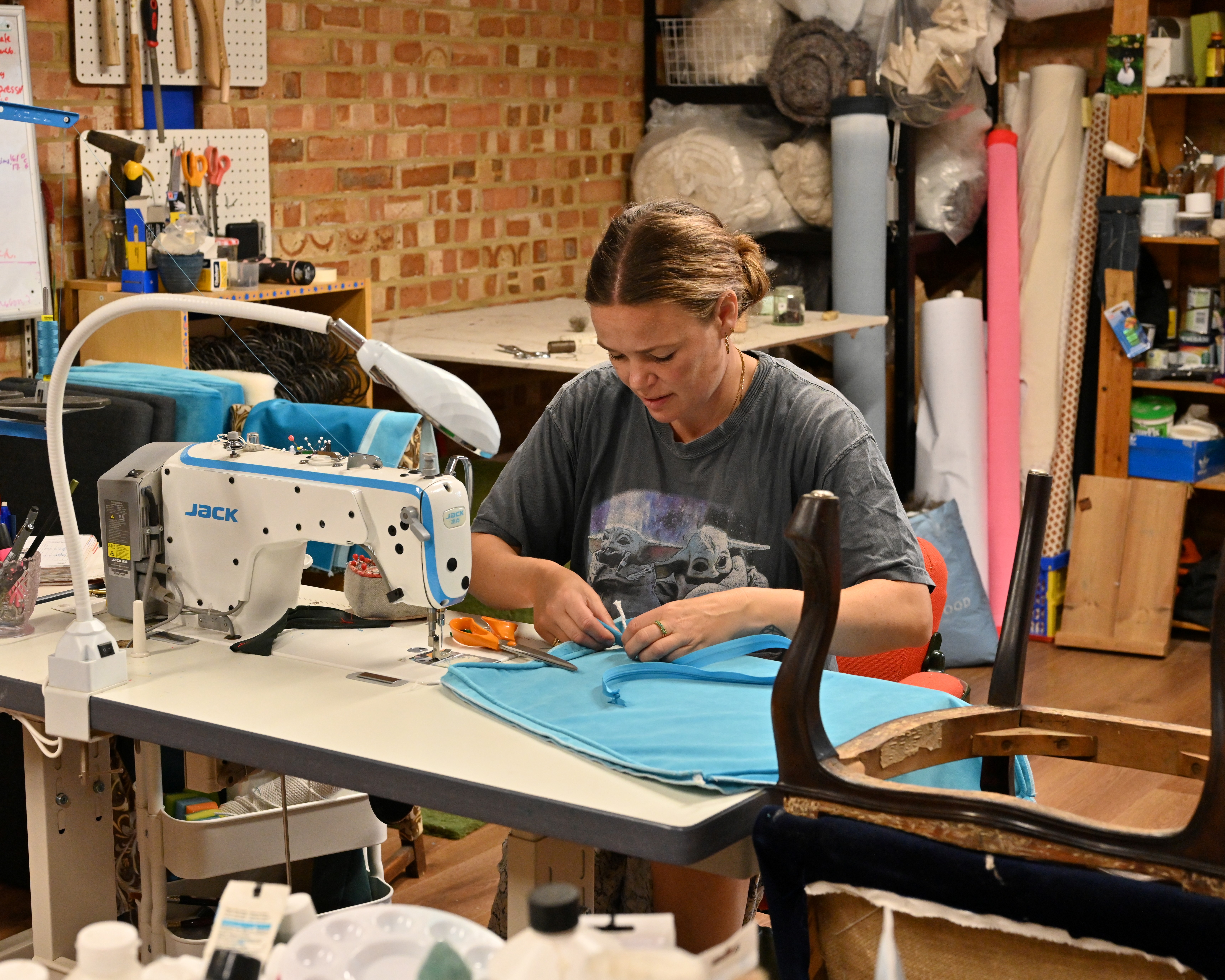 An female upholsterer photographed at work, up close and in portraits, in her studio, filled with wooden furniture, colorful fabric rolls, and mockup designs, and illuminated by bright light.
