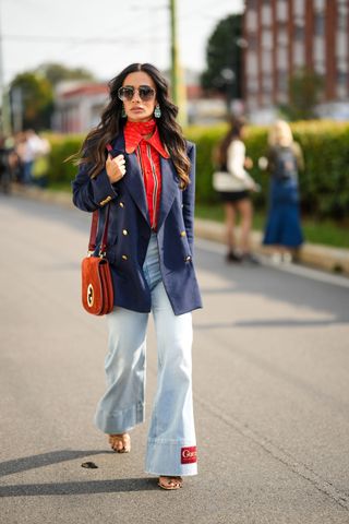 A guest wears sunglasses, silver and crystal earrings, a red with white GG monogram print pattern silk scarf from Gucci, a red with black and white striped print pattern shirt, a navy blue with gold buttoned blazer jacket, a camel suede shoulder bag from Gucci, blue faded denim wide legs pants from Gucci, gold shiny leather strappy heels sandals , outside Gucci, during the Milan Fashion Week - Womenswear Spring/Summer 2023 on September 23, 2022 in Milan, Italy.