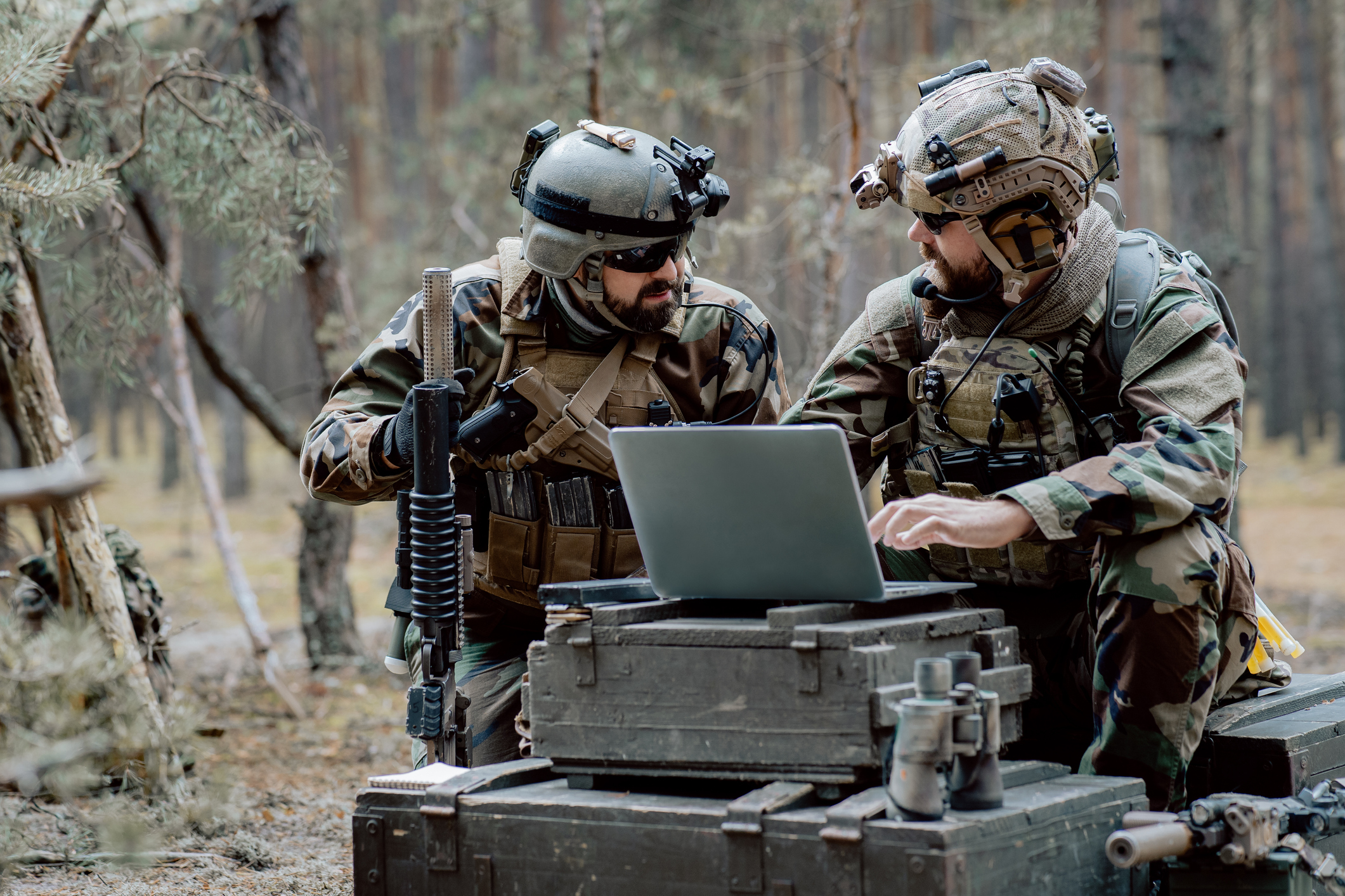 Bearded soldiers in uniform sit on military transport crates, analyze data on a laptop and work out tactics at a temporary forest base. In the background, you can see a soldier protecting the base.