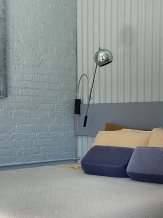 Grey bedroom with bed next to wall, dark grey headboard with lamp on wall above it.