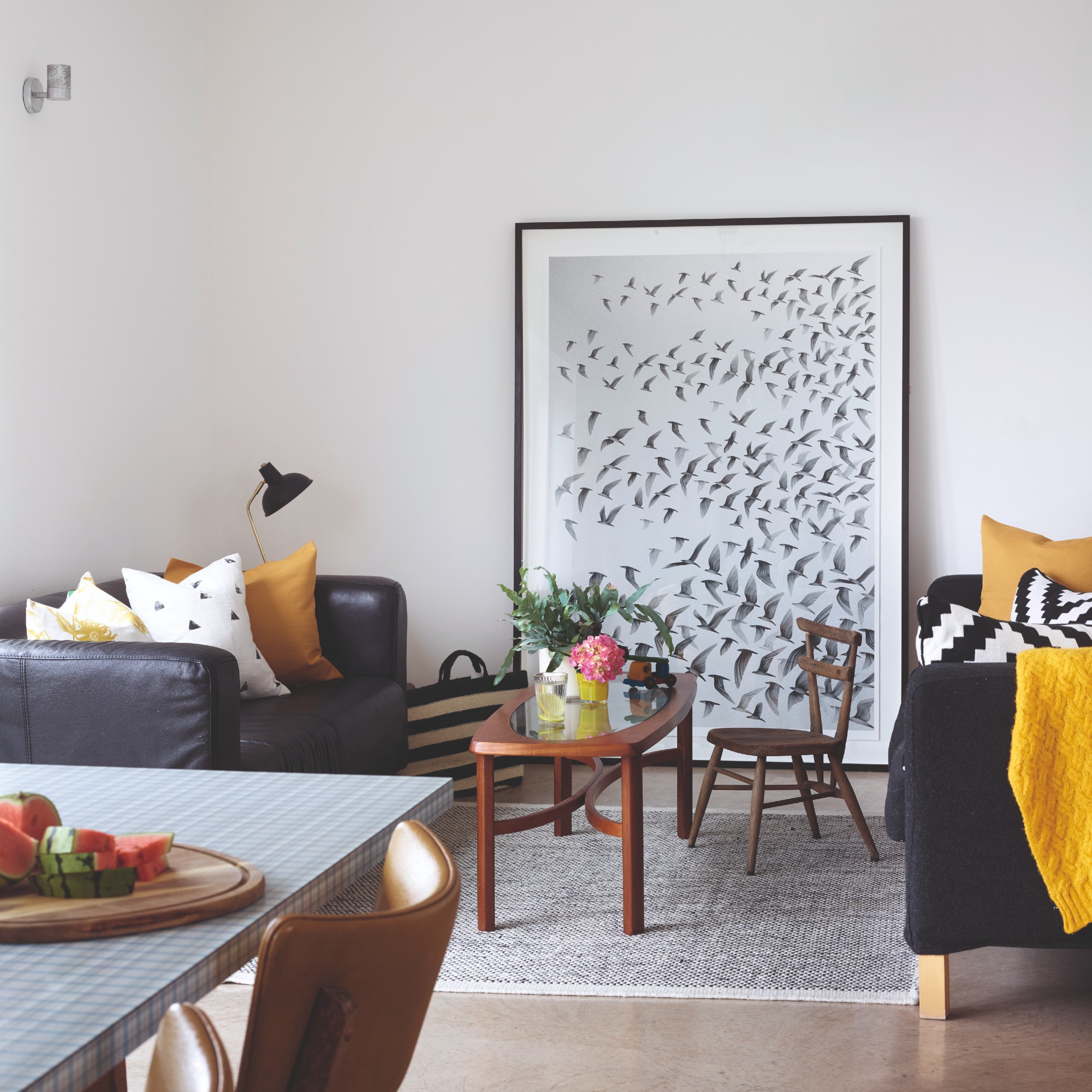 A living room with two black sofas, a dark wood and glass coffee table and a large framed art print leaning against the wall