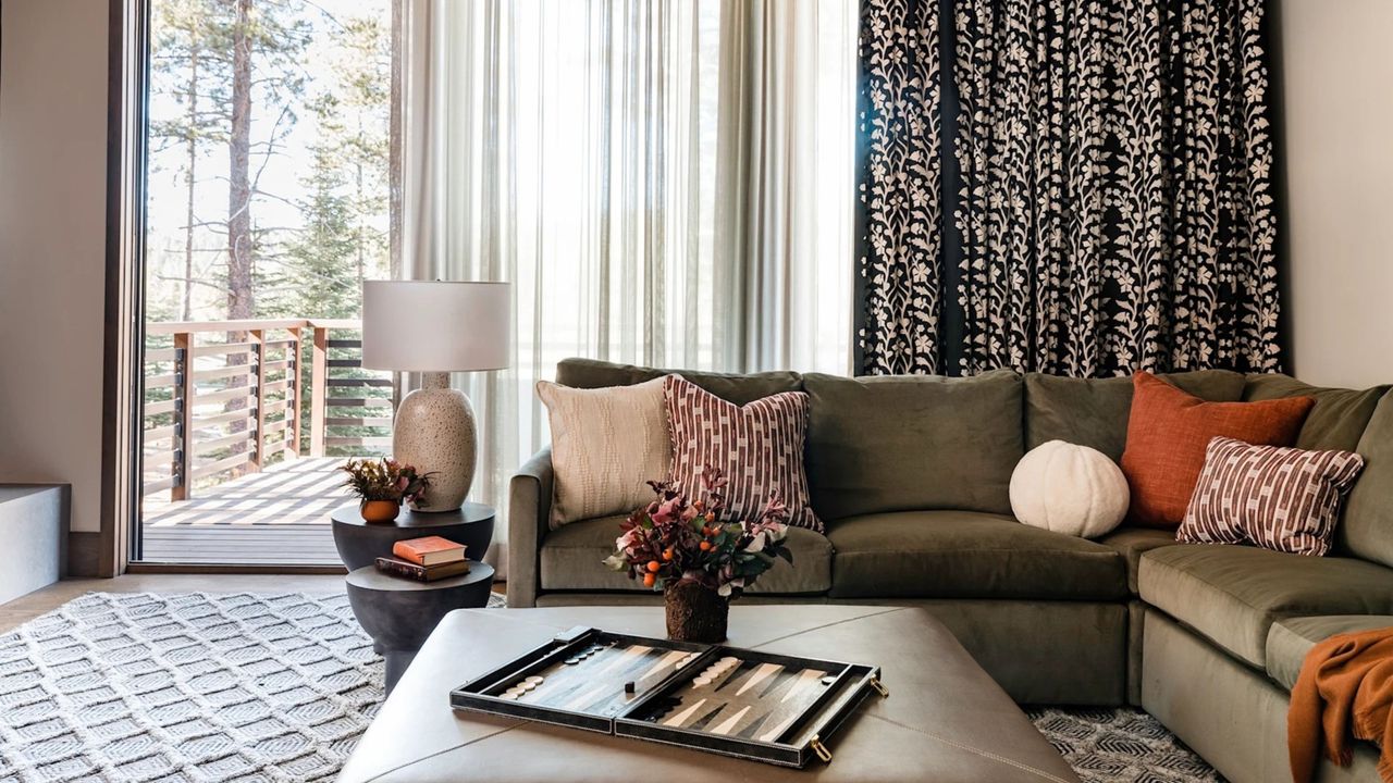 living room with patterned curtains hung over sheers, green l-shaped sofa with patterned cushions, leather ottoman coffee table with backgammon board and vase of flowers on it
