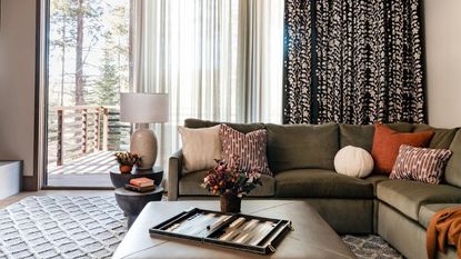 living room with patterned curtains hung over sheers, green l-shaped sofa with patterned cushions, leather ottoman coffee table with backgammon board and vase of flowers on it