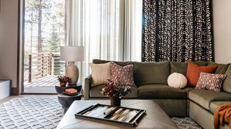 living room with patterned curtains hung over sheers, green l-shaped sofa with patterned cushions, leather ottoman coffee table with backgammon board and vase of flowers on it