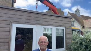 A man standing in front of his garden shed with a crane in the background