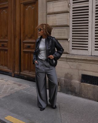 A woman wears gray trousers with a leather jacket on a sidewalk in Paris.