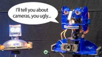 Talking Ohbot robots with speech bubble. One robot says "I'll tell you about cameras, you ugly..." to the other.