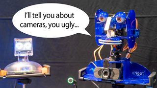 Talking Ohbot robots with speech bubble. One robot says "I'll tell you about cameras, you ugly..." to the other.