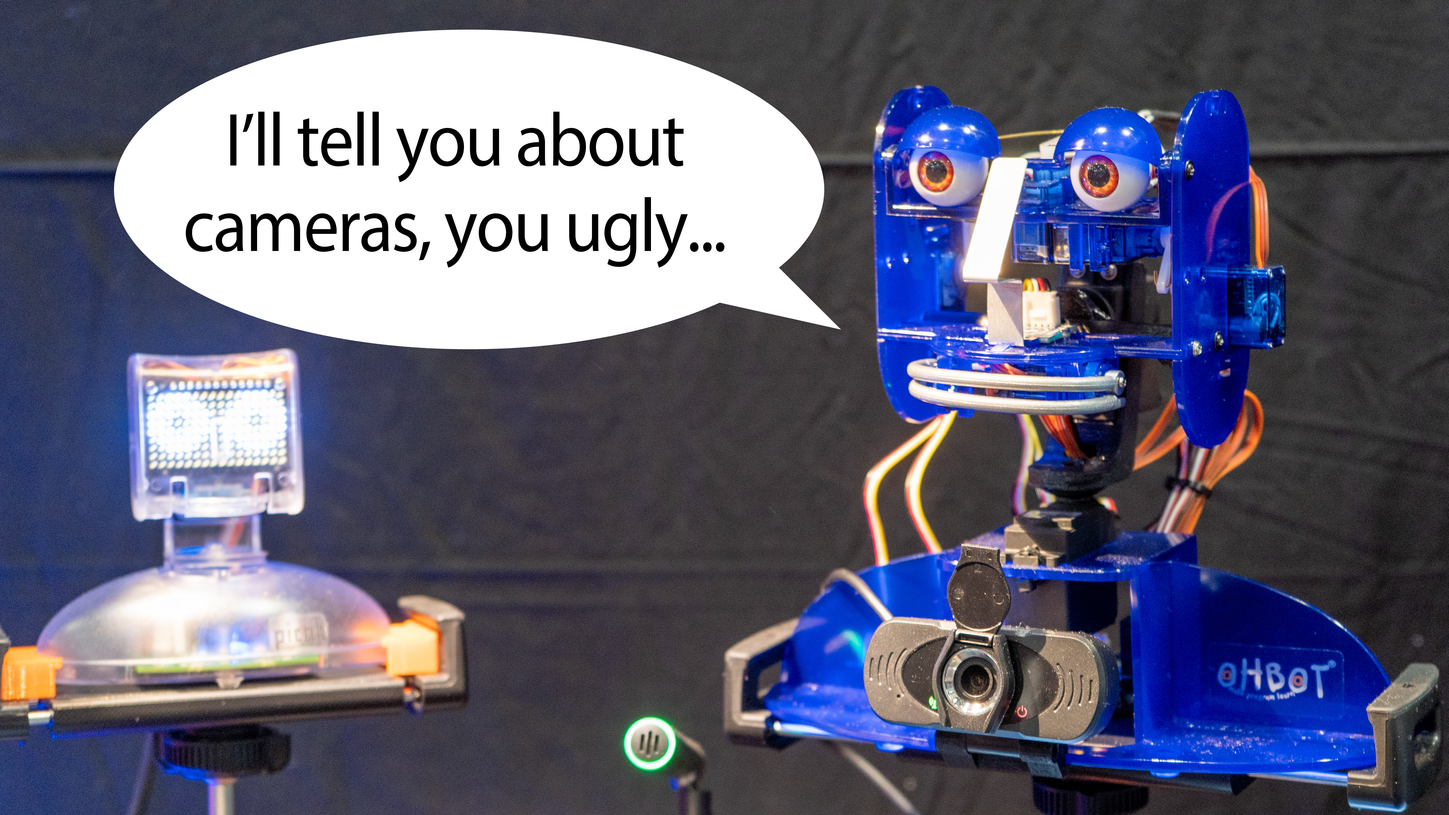 Talking Ohbot robots with speech bubble. One robot says "I'll tell you about cameras, you ugly..." to the other.