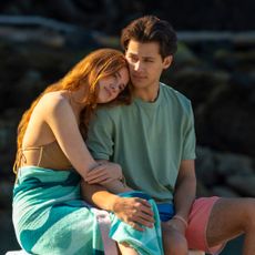 a young woman with a towel wrapped around her waist rests her head on a young man's shoulder at the beach at sunset in a still from every year after