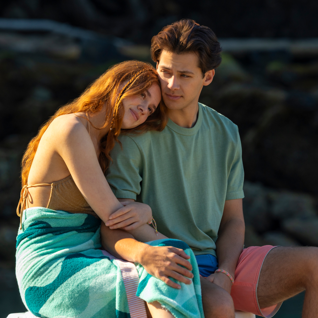 a young woman with a towel wrapped around her waist rests her head on a young man's shoulder at the beach at sunset in a still from every year after