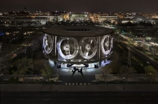 Hirshhorn Museum and Sculpture Garden photographed from high ground at night. The building is circular with a projection of auto wheels on the walls.