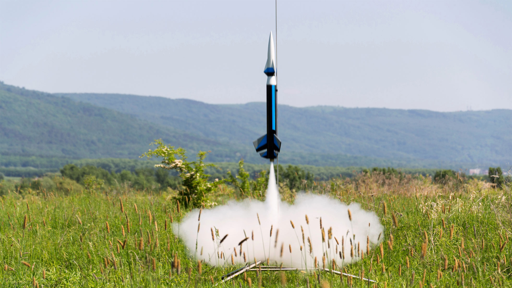 A blue model rocket with a white pointy tip is launching vertically as white vapor is produced at the bottom on a green grassy field with hills in the background.