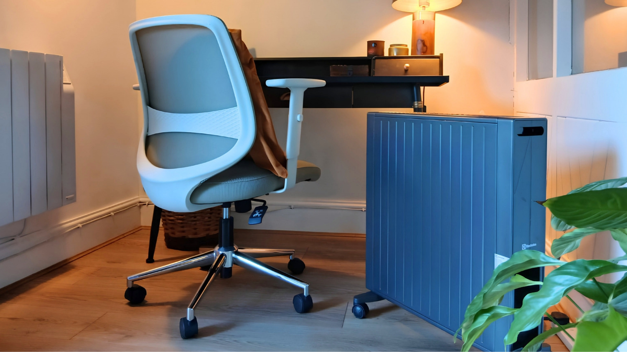 Home office with a grey mesh desk chair, wall radiator, blue portable heater, floating desk, and lamp.