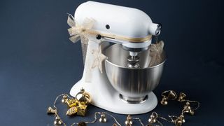 A white stand mixer wrapped in a gold bow surrounded by gold holiday decorations