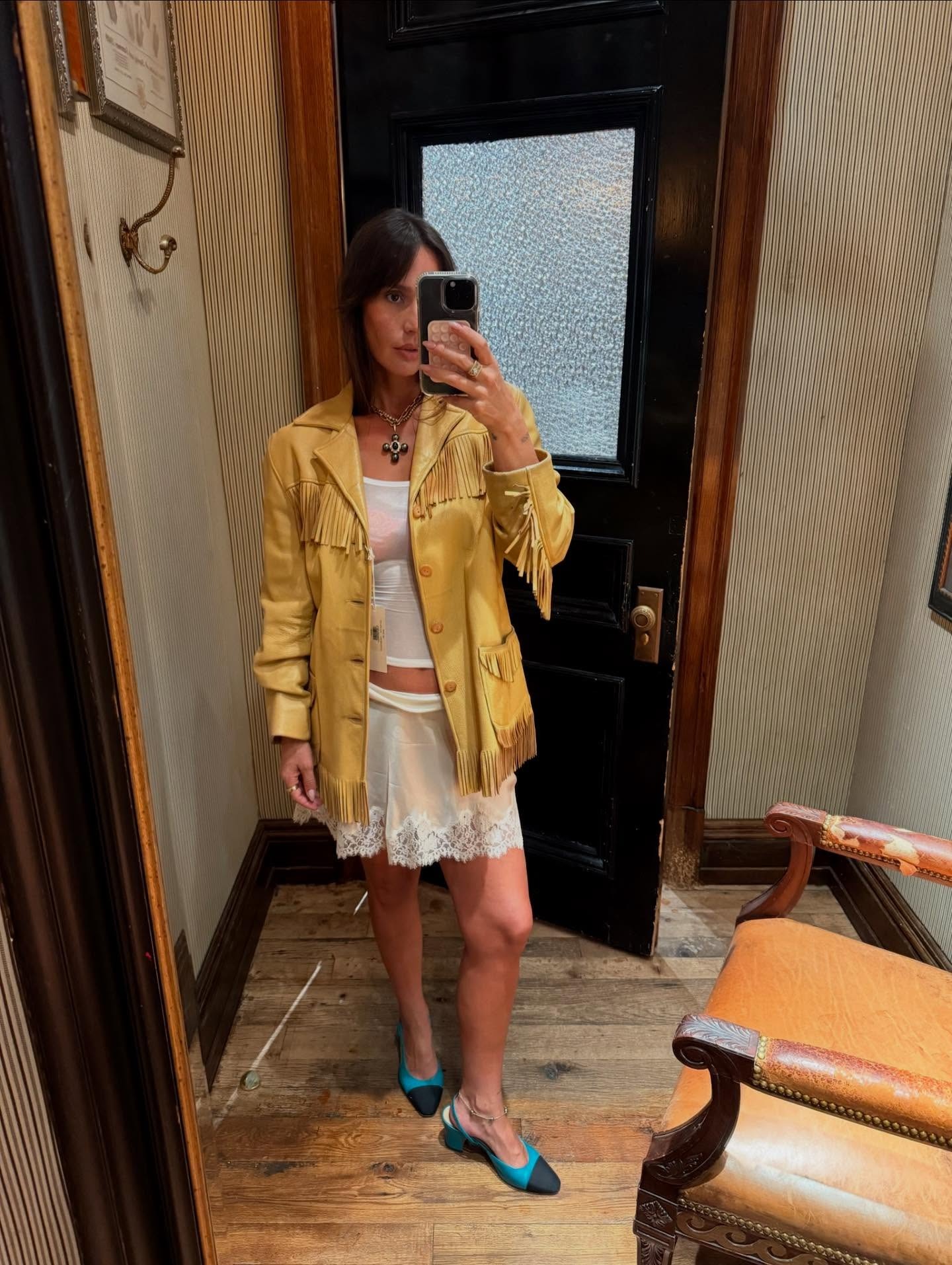Pia Baroncini takes a selfie in a light tan leather fringed jacket, sheer white t-shirt and slip skirt and blue Chanel heels.