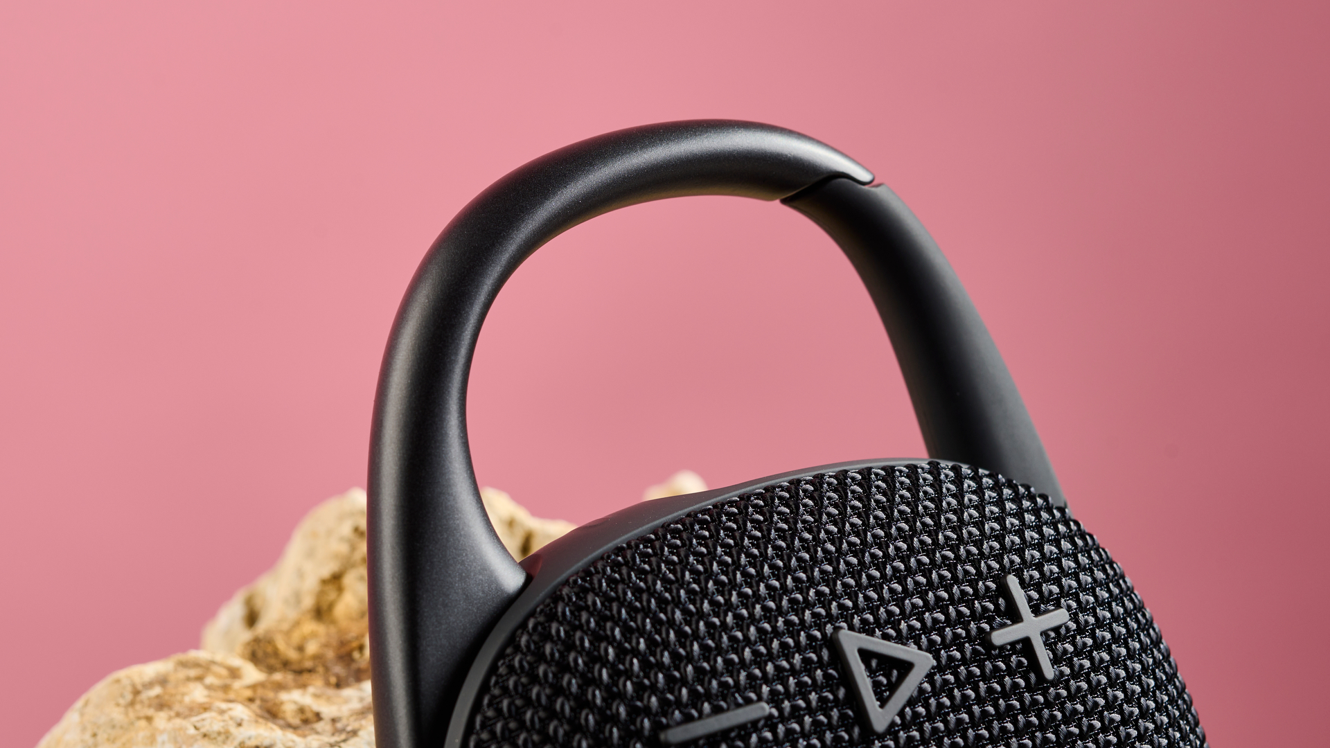 A close up of the carabiner on the top of the JBL Clip 5, pictured leaning against a sand coloured rock against a pink background,